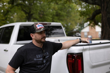 A man in a Bull Spurs Tee-Black Frost and cap leans against a white truck, holding a can of Coors Light. Trees and a brick house are visible in the background.