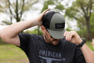 A person wearing a black and green “Rural” cap, sunglasses, and the Bull Spurs Tee in Black Frost stands outdoors, holding and adjusting the brim of their cap with one hand. Trees and grass are visible in the blurred background.