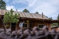 A man stands on the porch of a rustic building with a corrugated metal roof and wooden beams. He is wearing a hat, glasses, camouflage pants, and a Rural Cloth Hunt Pullover in Athletic Gray. A yellow sign on the porch reads 