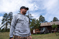 A man wearing a Rural Cloth Hunt Pullover-Athletic Gray hoodie and camouflage pants stands outdoors with trees and a rustic building in the background. He is wearing a black cap and a watch, looking towards his right. The sky is partly cloudy, setting the perfect scene for hunting enthusiasts.