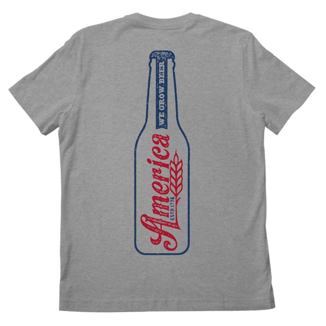 The American Cattle Co Longneck Tee is a gray shirt featuring a large blue beer bottle outline on the back, “America” in red script, a wheat graphic, and “We Grow Beer” printed vertically—ideal for beer lovers.