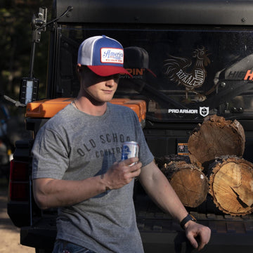 A man in an American Cattle Co Old School Country - Heather Gray T-shirt and a "Make America Cowboy Again" hat leans against a truck with Pabst Blue Ribbon in hand, firewood stacked on the tailgate.