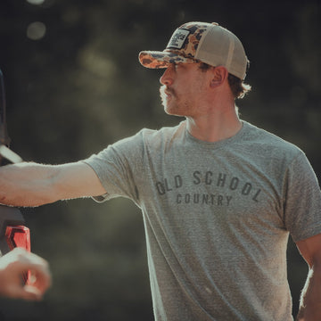 A man stands outdoors in sunlight, wearing an American Cattle Co Old School Country - Heather Gray T-shirt and a camouflage cap, his arm resting on a vehicle. This ultra-soft classic tee is proudly made in the USA. The background is blurred.