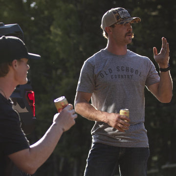 Two men stand outdoors near a vehicle, both in caps and casual wear. One wears American Cattle Co's ultra-soft tee in heather gray; the other sports the Old School Country - Heather Gray tee. Sunlight and trees fill the background as they each hold a drink.