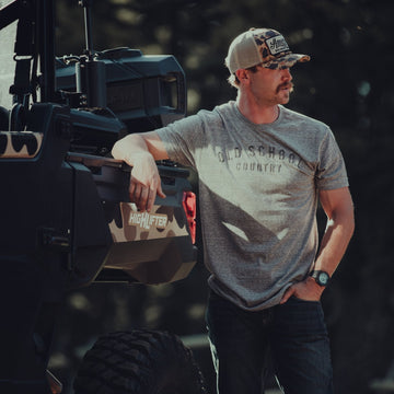 A man in a camouflage hat, sunglasses, and an American Cattle Co Old School Country - Heather Gray tee leans against an off-road vehicle outdoors, with softly blurred trees in the background.