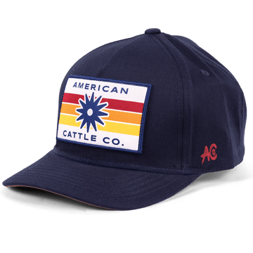 Introducing the Retro Spur Hat: a navy blue snapback ranch hat with a rectangular patch on the front showcasing colorful stripes, a starburst design, and “American Cattle Co.” text. A small red “AC” logo is stitched on the side, completing this classic western look.