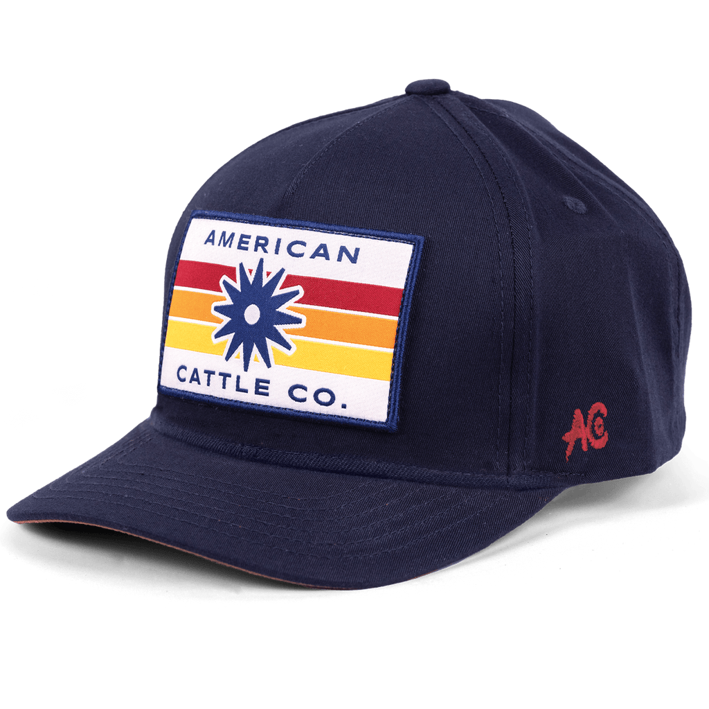 Introducing the Retro Spur Hat: a navy blue snapback ranch hat with a rectangular patch on the front showcasing colorful stripes, a starburst design, and “American Cattle Co.” text. A small red “AC” logo is stitched on the side, completing this classic western look.