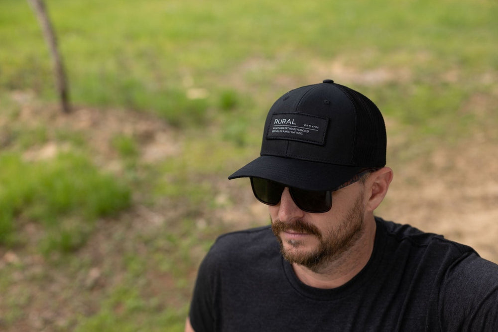 A bearded man wearing the American Cattle Co Rural Def Hat and black sunglasses stands outdoors in a dark t-shirt, with green grass and a dirt path behind him.
