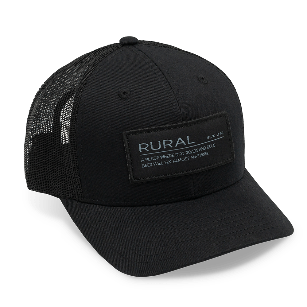 The American Cattle Co Rural Def Hat is a black cap with a breathable mesh back, adjustable snapback, and a rectangular patch reading: "RURAL EST. 1776. A PLACE WHERE DIRT ROADS AND COLD BEER WILL FIX ALMOST ANYTHING.