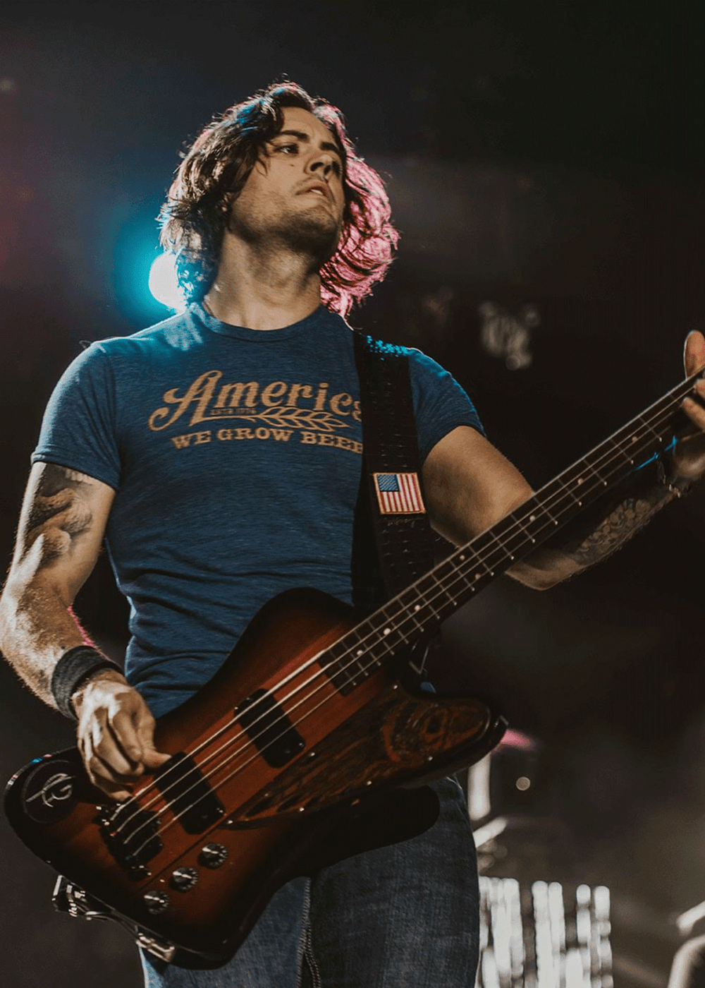 On stage, a long-haired musician plays electric bass in jeans and the Rural Cloth We Grow Beer Tee - Royal, with stage lights glowing behind.