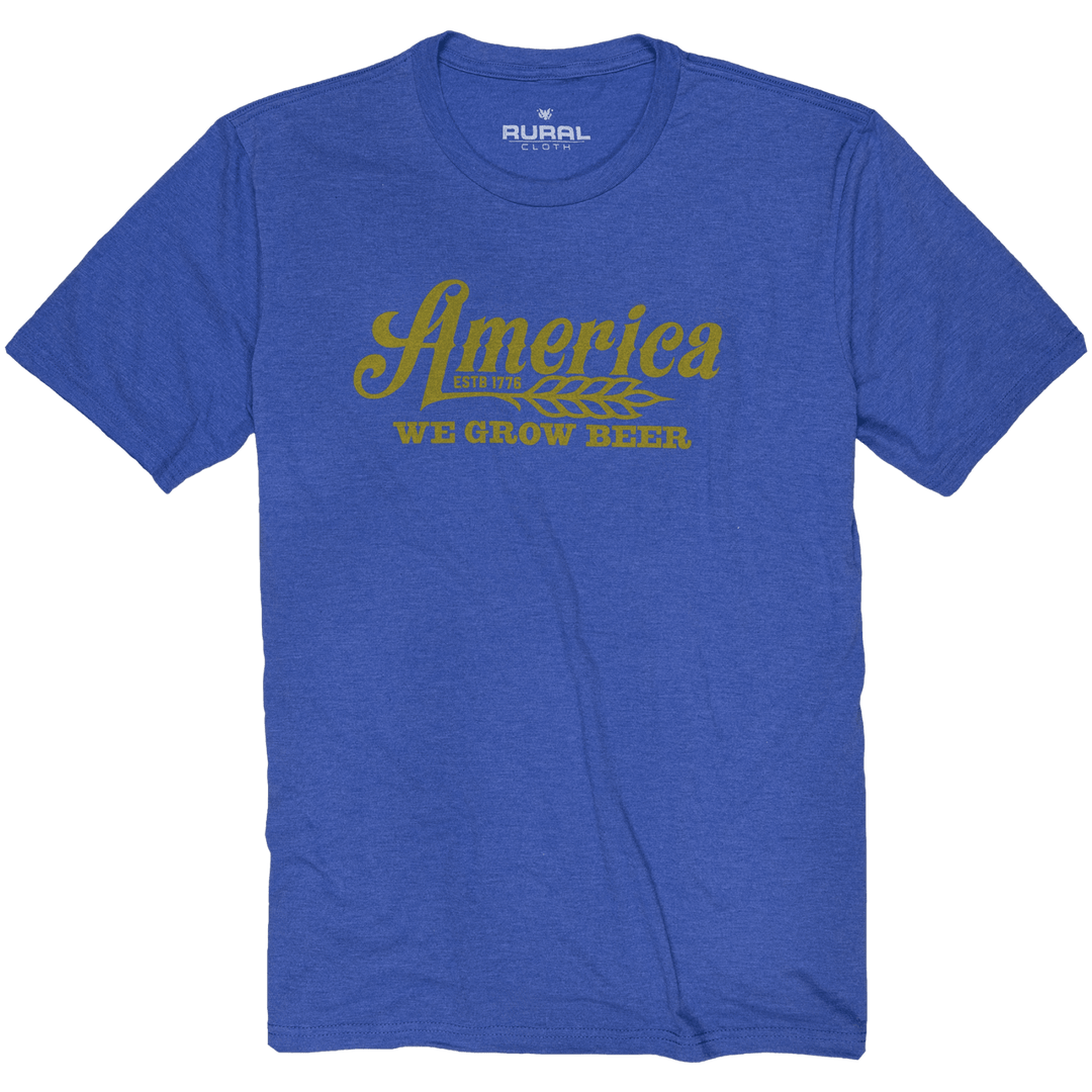 The We Grow Beer Tee - Royal by Rural Cloth features yellow "America Estd 1776 WE GROW BEER" text and a wheat stalk graphic on blue premium fabric, offering a modern athletic fit.