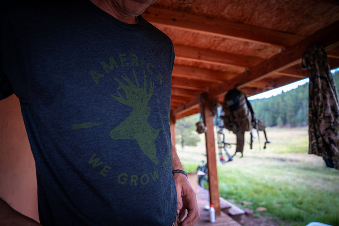 Wearing the American Cattle Co We Grow Deer Tee-Black with its bold deer graphic and "AMERICA WE GROW," a person stands under a wooden porch with hunting gear, embracing wildlife stewardship against a grassy landscape backdrop.