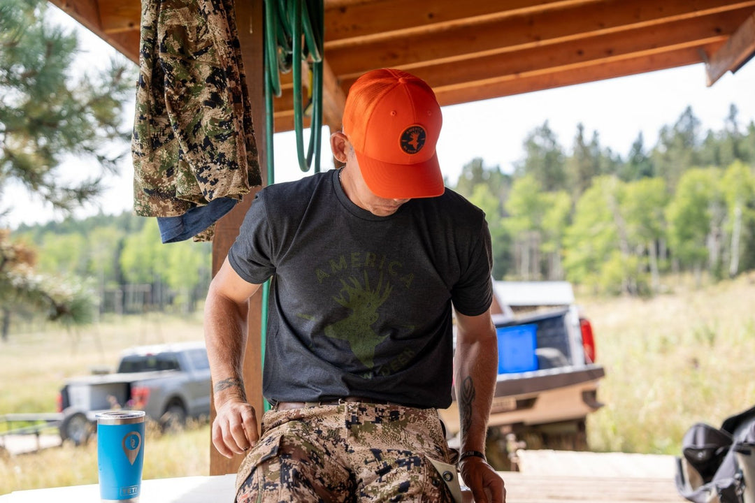 Wearing an American Cattle Co We Grow Deer Tee-Black, an orange cap, and camo pants, a person sits under a wooden shelter. A vehicle and forest fill the background; a camo jacket and blue cup nearby show their passion for wildlife stewardship.