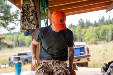 Wearing an American Cattle Co We Grow Deer Tee-Black, an orange cap, and camo pants, a person sits under a wooden shelter. A vehicle and forest fill the background; a camo jacket and blue cup nearby show their passion for wildlife stewardship.