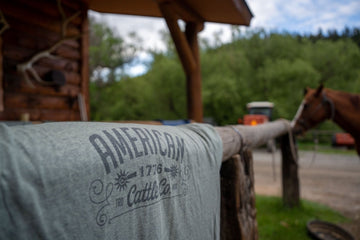 A gray Western Tee from American Cattle Co with “AMERICAN 1776 Cattle Co.” printed on it hangs over a wooden fence, with a brown horse, red vehicle, log cabin, and trees in the background—reflecting true American cattle spirit.